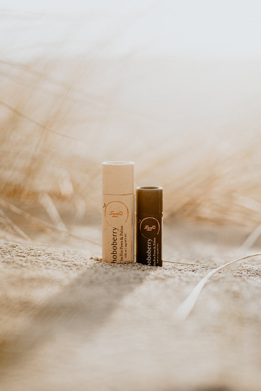 Two hoboberry balms. One beige kraft paper tube standing next to black kraft paper tube in the sand 