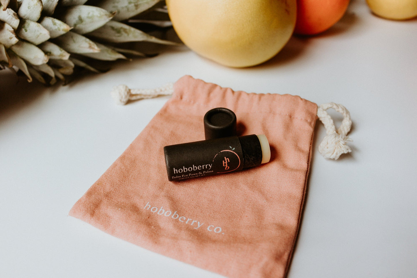 kraft paper tube balm for paws and palms laying on pink cotton draw string bag with fruits lined up in the back