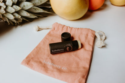 kraft paper tube balm for paws and palms laying on pink cotton draw string bag with fruits lined up in the back