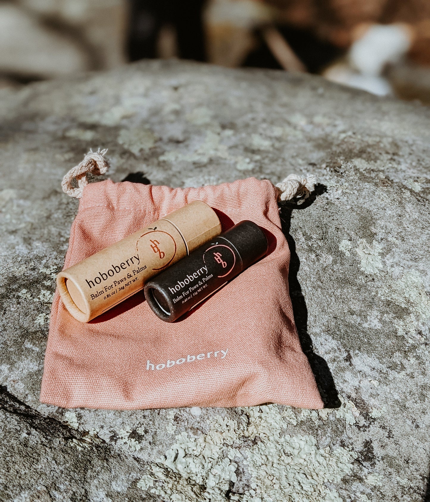 Two hoboberry balms. One beige kraft paper tube and one black kraft paper tube both laying down on pink cotton draw string bag outside on top of a rock